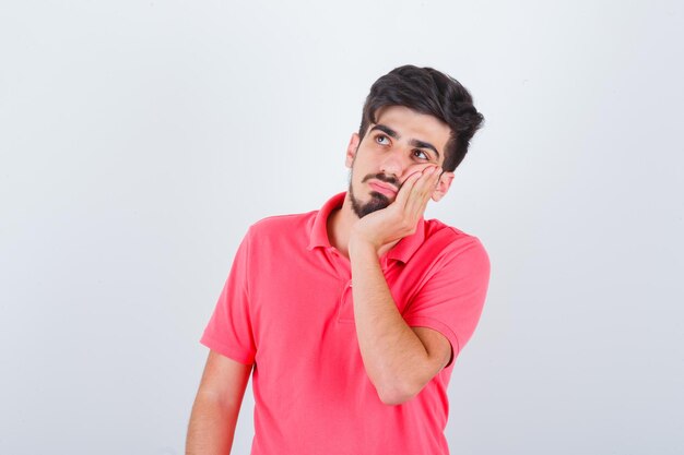 Young male leaning cheek on palm in pink t-shirt and looking pensive. front view.