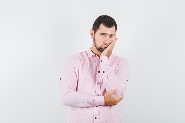 Молодой мужчина в розовой рубашке прислонился щекой к поднятой ладони и выглядит усталым