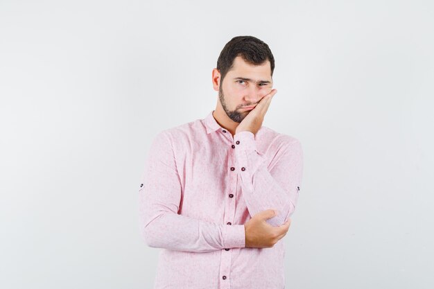 Young male leaning cheek on raised palm in pink shirt and looking fatigued