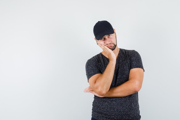 Young male leaning cheek on raised palm in t-shirt and cap and looking tired