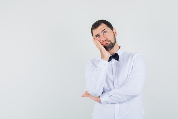 Young male leaning cheek on raised palm in white shirt and looking pensive. front view.