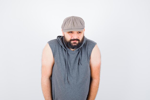 Young male looking away in sleeveless hoodie, cap and looking bored , front view.