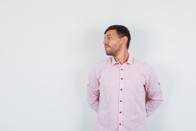 Молодой мужчина смотрит в сторону с руками на спине в розовой рубашке и смотрит задумчиво, вид спереди.