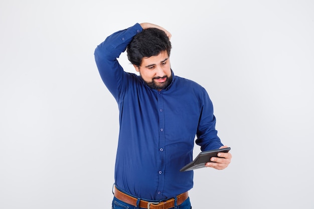 Young male looking at calculator while holding hand on head in royal blue shirt front view.