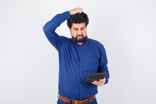 Young male looking at calculator while scratching his head in royal blue shirt front view.