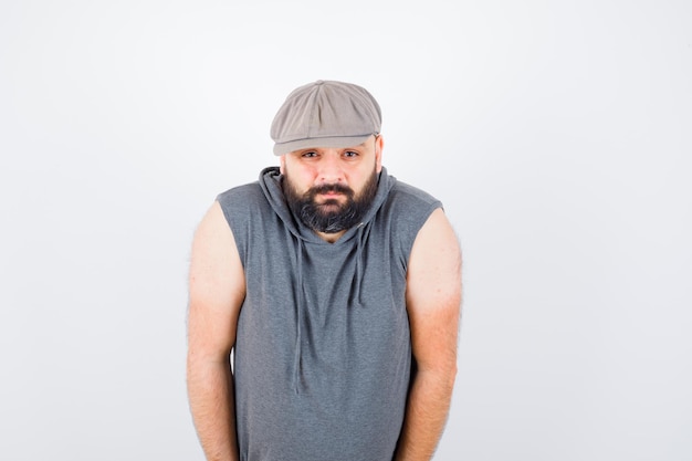 Free photo young male looking at camera in sleeveless hoodie, cap and looking bored. front view.