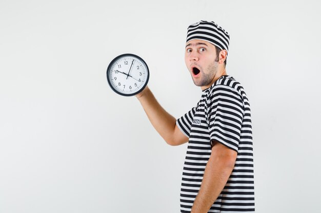 Young male looking at clock in striped t-shirt,cap and looking surprised. .