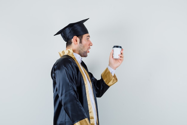 Young male looking at cup of coffee in graduate uniform and looking surprised. .