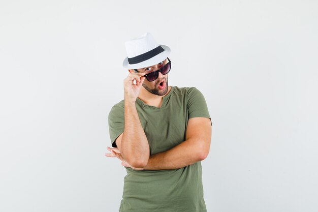 Young male looking over glasses in green t-shirt and hat and looking curious