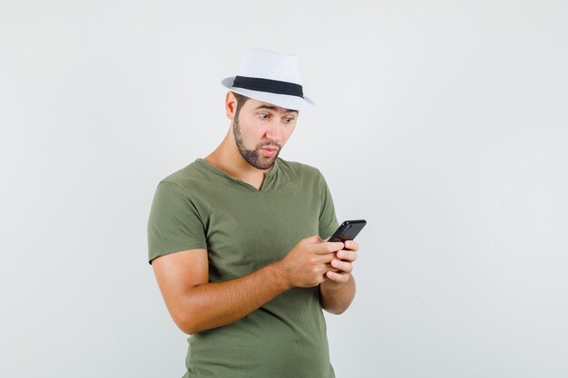 Молодой мужчина смотрит на мобильный телефон в зеленой футболке и шляпе и выглядит изумленным