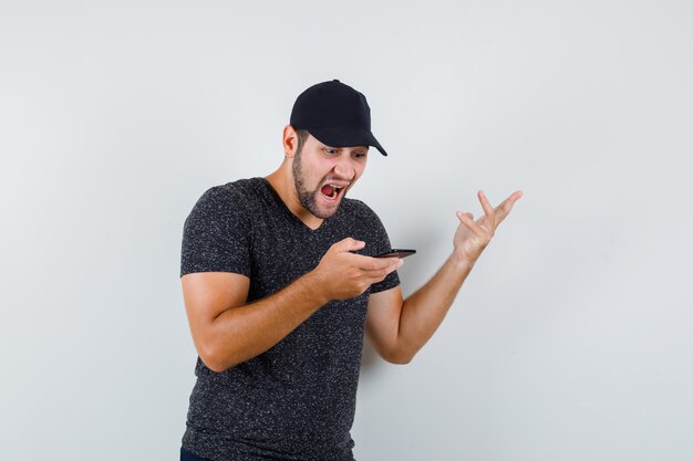 Young male looking at mobile phone in t-shirt and cap, jeans and looking angry