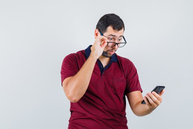 Young male looking at phone in red t-shirt,glasses and looking surprised. front view.