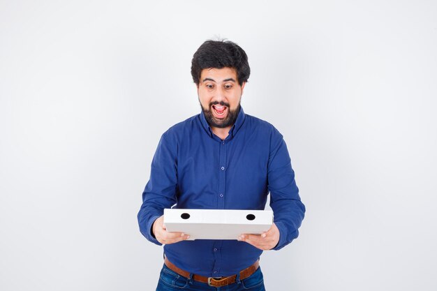 Young male looking at pizza box in shirt, jeans and looking joyful. front view.