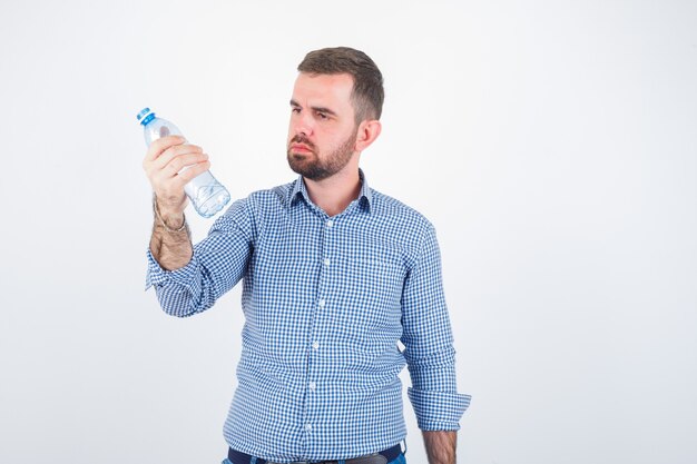 Молодой мужчина смотрит на пластиковую бутылку с водой в рубашке, джинсах и смотрит вдумчивый, вид спереди.