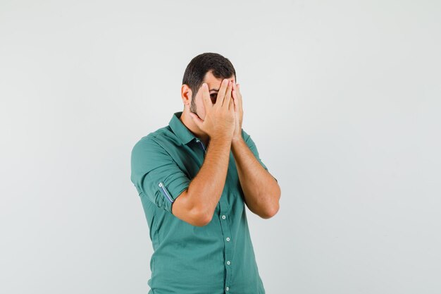 Young male looking through fingers in green shirt and looking worried , front view.