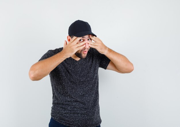 Young male looking through fingers in t-shirt and cap, jeans and looking curious