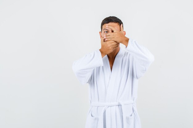 Young male looking through fingers while covering mouth in white bathrobe and looking scared. front view.