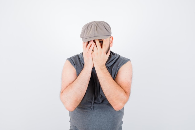 Free photo young male looking through fingers with one eye in sleeveless hoodie, cap and looking cute , front view.