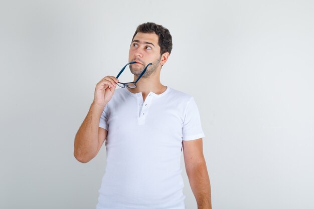 Young male looking up and holding glasses in white t-shirt and looking thoughtful