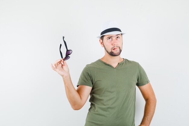 Young male looking up while holding glasses in green t-shirt and hat and looking hesitant