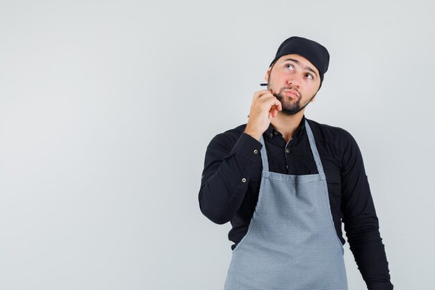 Young male looking up with finger on jaw in shirt, apron and looking pensive. front view.