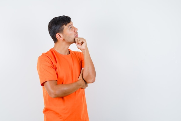 Young male looking up with hand on chin in orange t-shirt and looking pensive