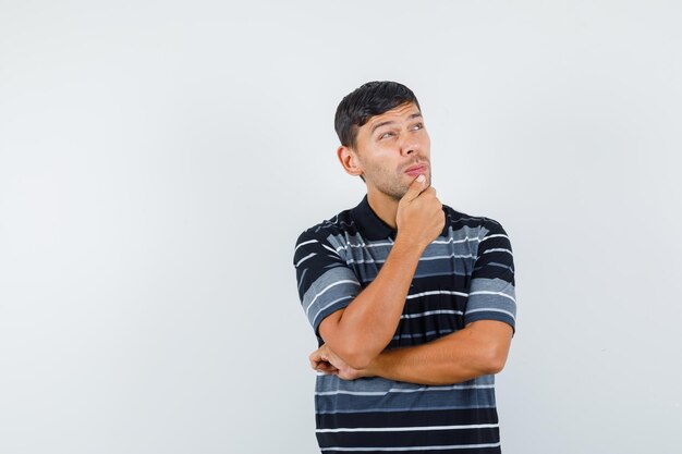 Молодой мужчина смотрит вверх с рукой на подбородке в футболке и смотрит задумчиво, вид спереди.