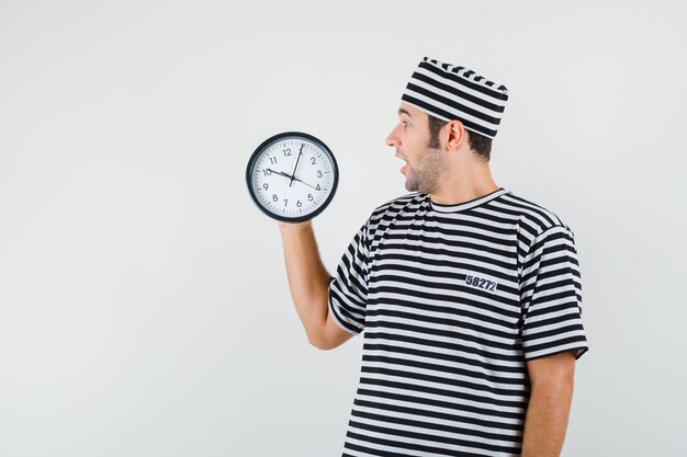 Young male looking at wall clock in t-shirt, hat and looking surprised , front view.