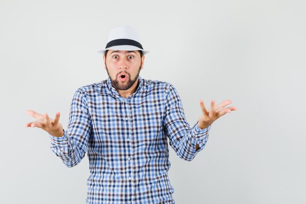 Young male making asking question gesture in checked shirt, hat and looking puzzled , front view.