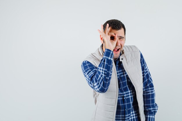 Young male making round on his eye in shirt,sleeveless jacket and looking amused. front view. space for text