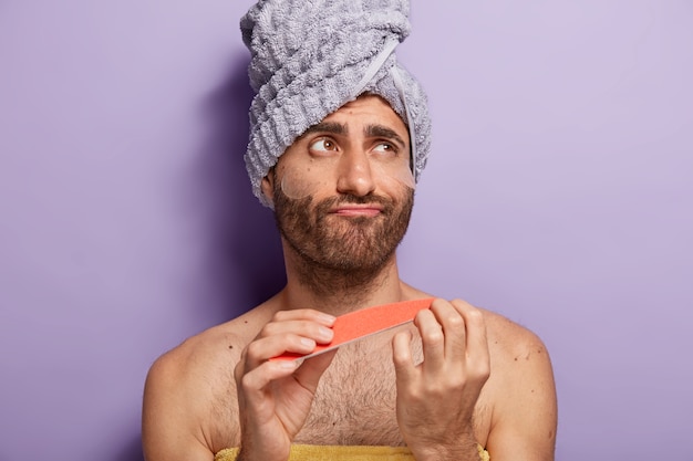 Young male model does manicure with nail file, wears silicone patches under eyes, has beauty treatments, wears towel on head, stands with bare torso against purple wall, looks aside pensively