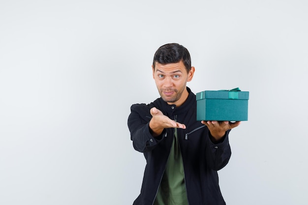 Young male offering gift box in t-shirt, jacket and looking merry , front view.