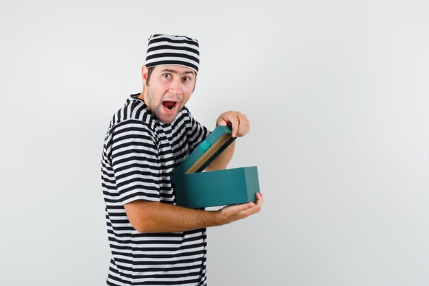 Young male opening gift box in t-shirt, hat and looking curious .