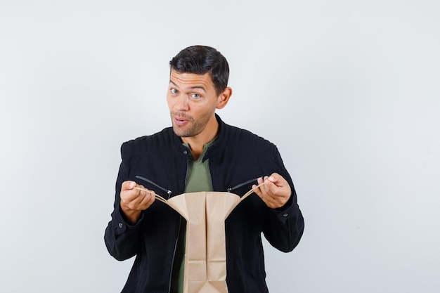 Free photo young male opening paper bag in t-shirt, jacket and looking impatient , front view.