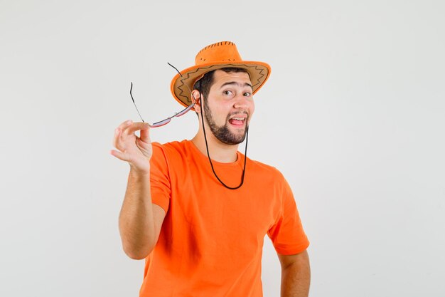 Молодой мужчина в оранжевой футболке, шляпе держит очки и выглядит весело, вид спереди.