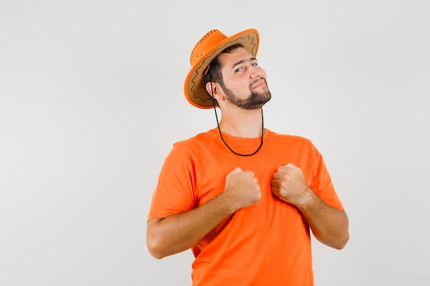 Young male in orange t-shirt, hat keeping clenched fists on chest and looking confident , front view.