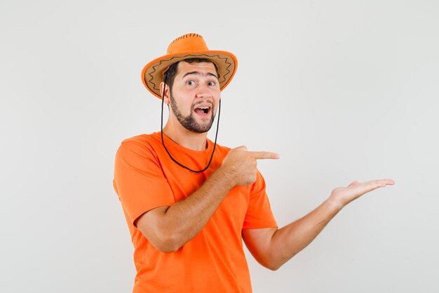 Молодой мужчина в оранжевой футболке, шляпа, указывающая на растопыренную ладонь, вид спереди.