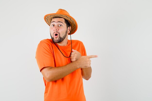 Young male in orange t-shirt, hat pointing to the side and looking amazed , front view.