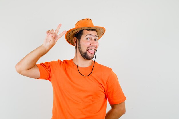 Молодой мужчина в оранжевой футболке, шляпе показывает знак победы, высунув язык и выглядит смешно, вид спереди.