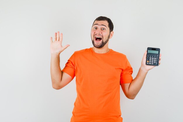 Young male in orange t-shirt holding calculator, showing palm and looking happy , front view.