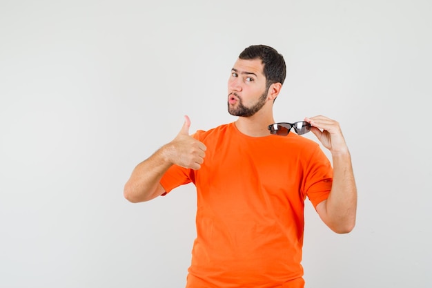 Young male in orange t-shirt holding sunglasses with thumb up and looking confident , front view.