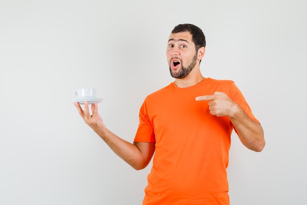 Young male in orange t-shirt pointing at cup with saucer and looking positive , front view.
