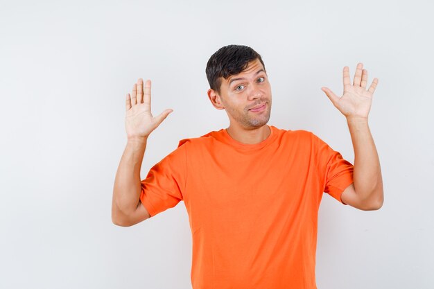 Молодой мужчина в оранжевой футболке поднимает ладони в жесте капитуляции и выглядит храбрым