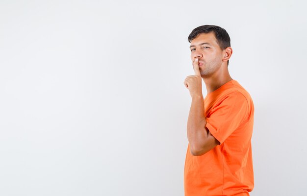 沈黙のジェスチャーを示し、注意深く見えるオレンジ色のTシャツの若い男性