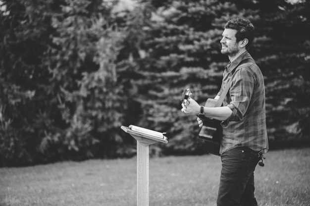 A young male in a park holding a guitar and playing a song from the Christian hymn book