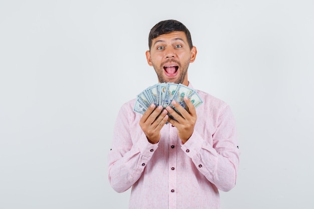 Young male in pink shirt holding dollar bills and looking cheerful , front view.