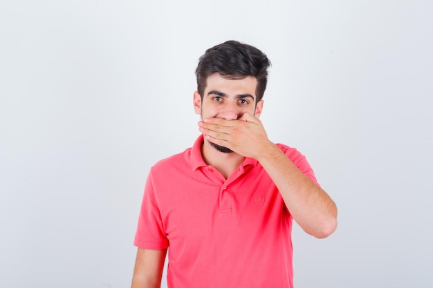 Young male in pink t-shirt holding hand on mouth and looking merry , front view.