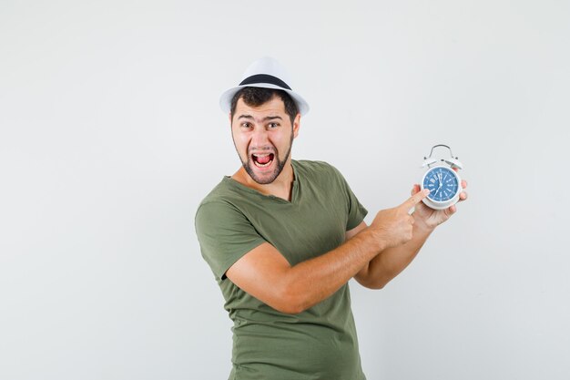 Free photo young male pointing at alarm clock in green t-shirt,hat and looking crazy