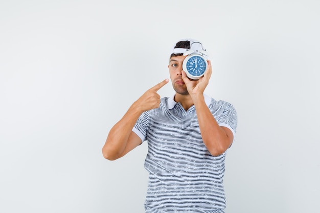 Free photo young male pointing at alarm clock in t-shirt and cap and looking serious