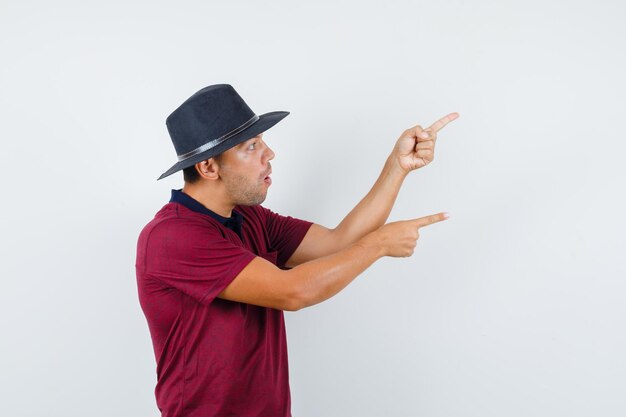 Young male pointing aside in red shirt,black hat and looking surprised. .